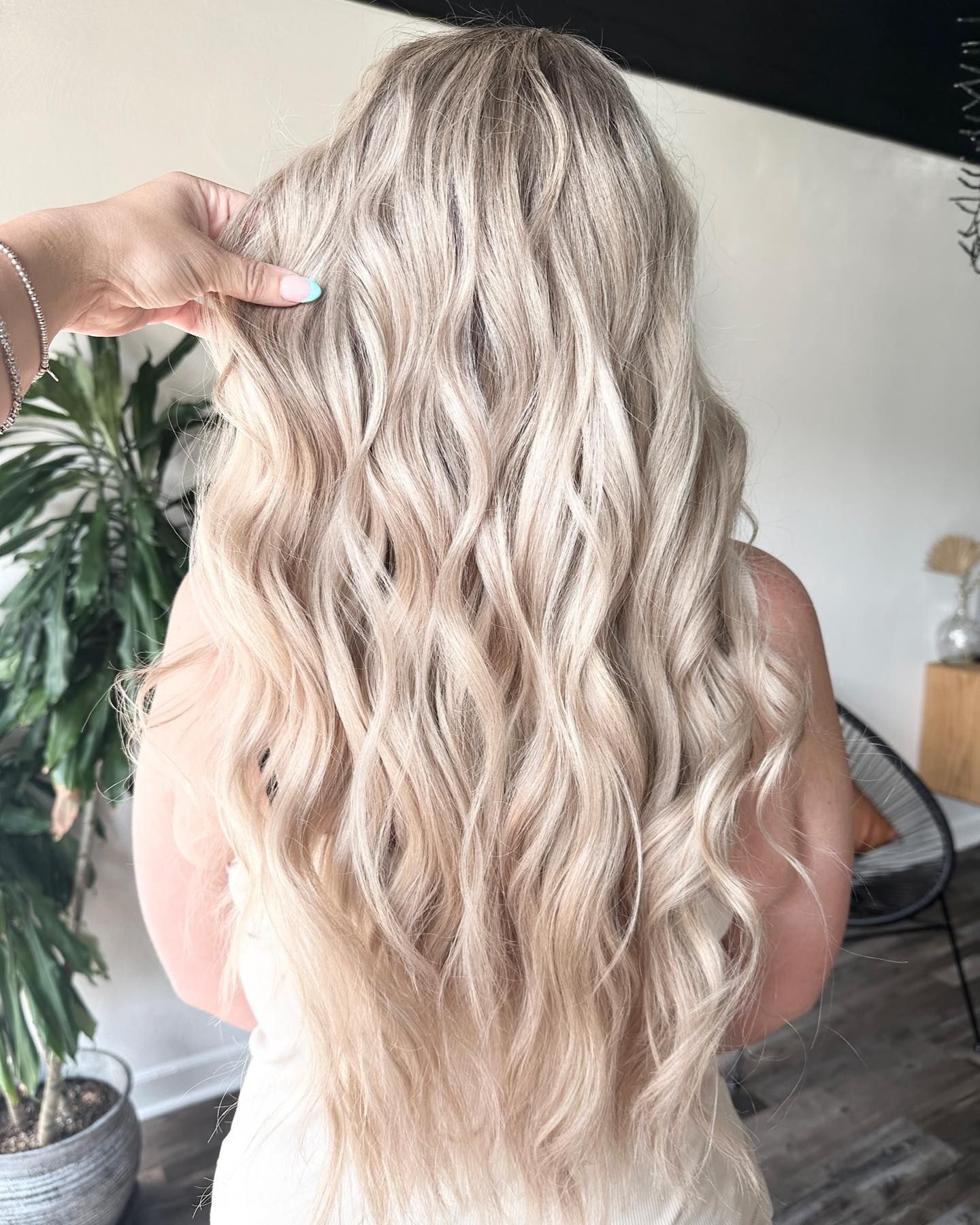 Long, wavy, light blonde hair. A hand lifts a section of the hair. Indoors, near a plant and chair.