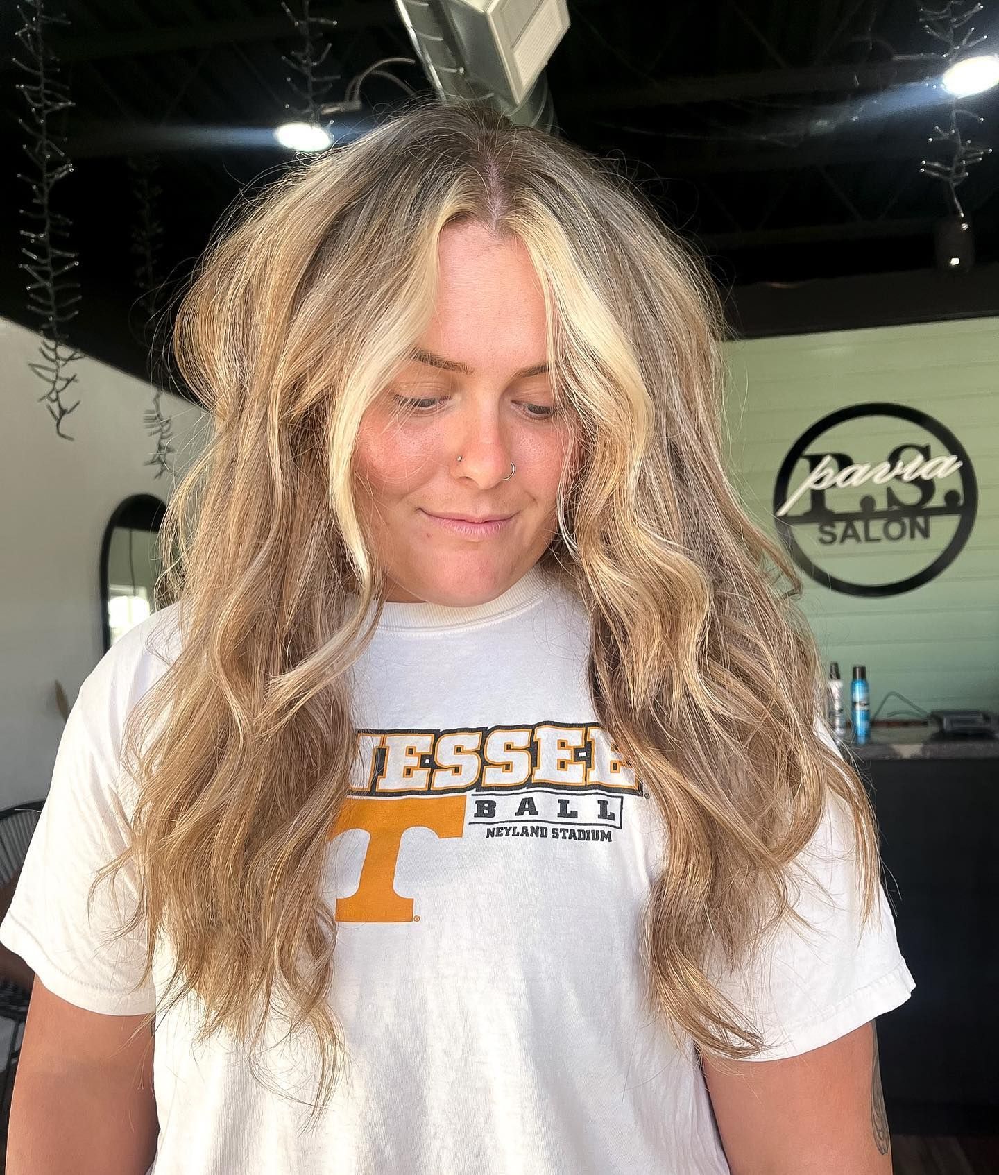 Woman with wavy blonde hair, wearing a white t-shirt, stands inside a salon.