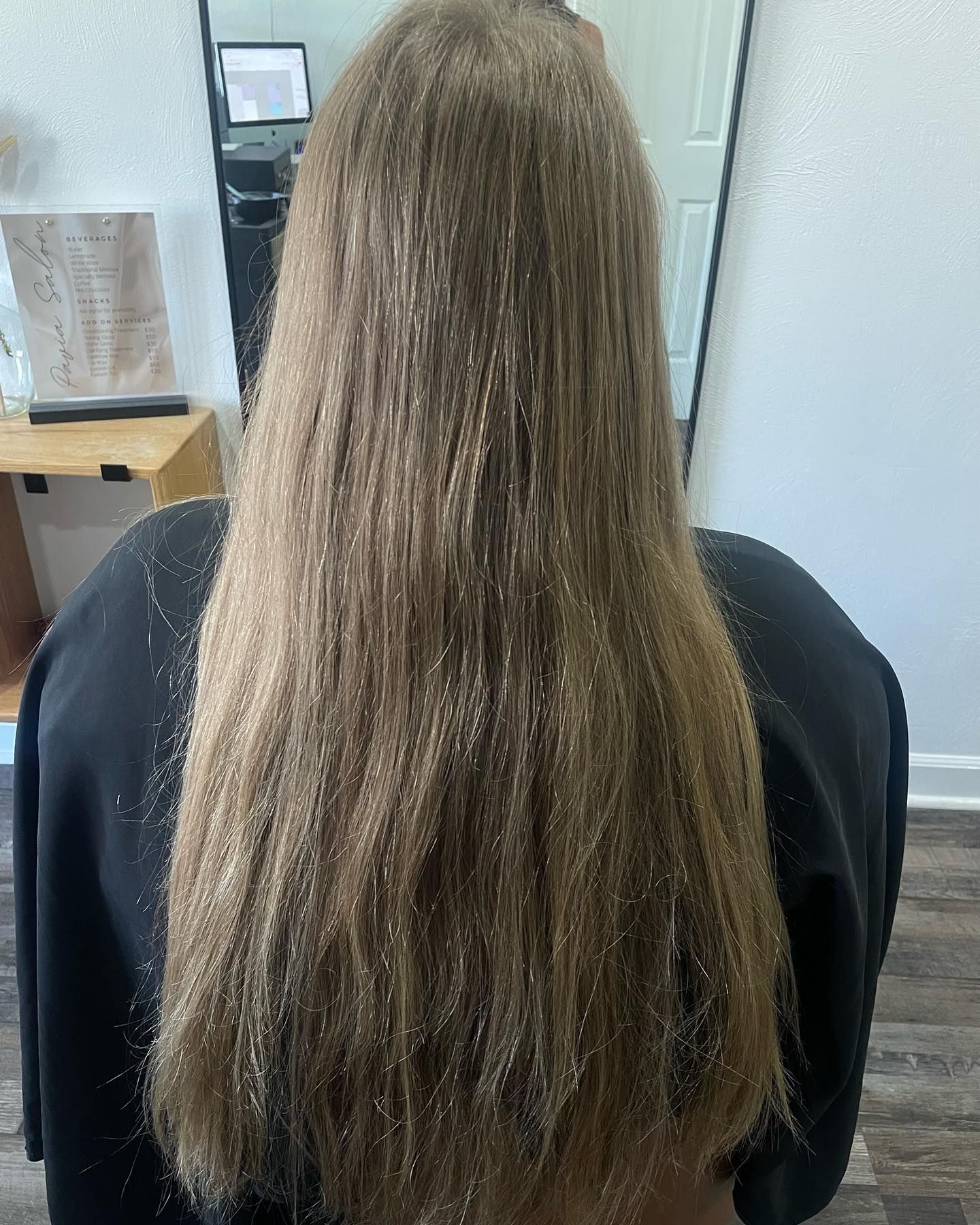 Long, light brown hair hangs down the back of a person, inside a salon.