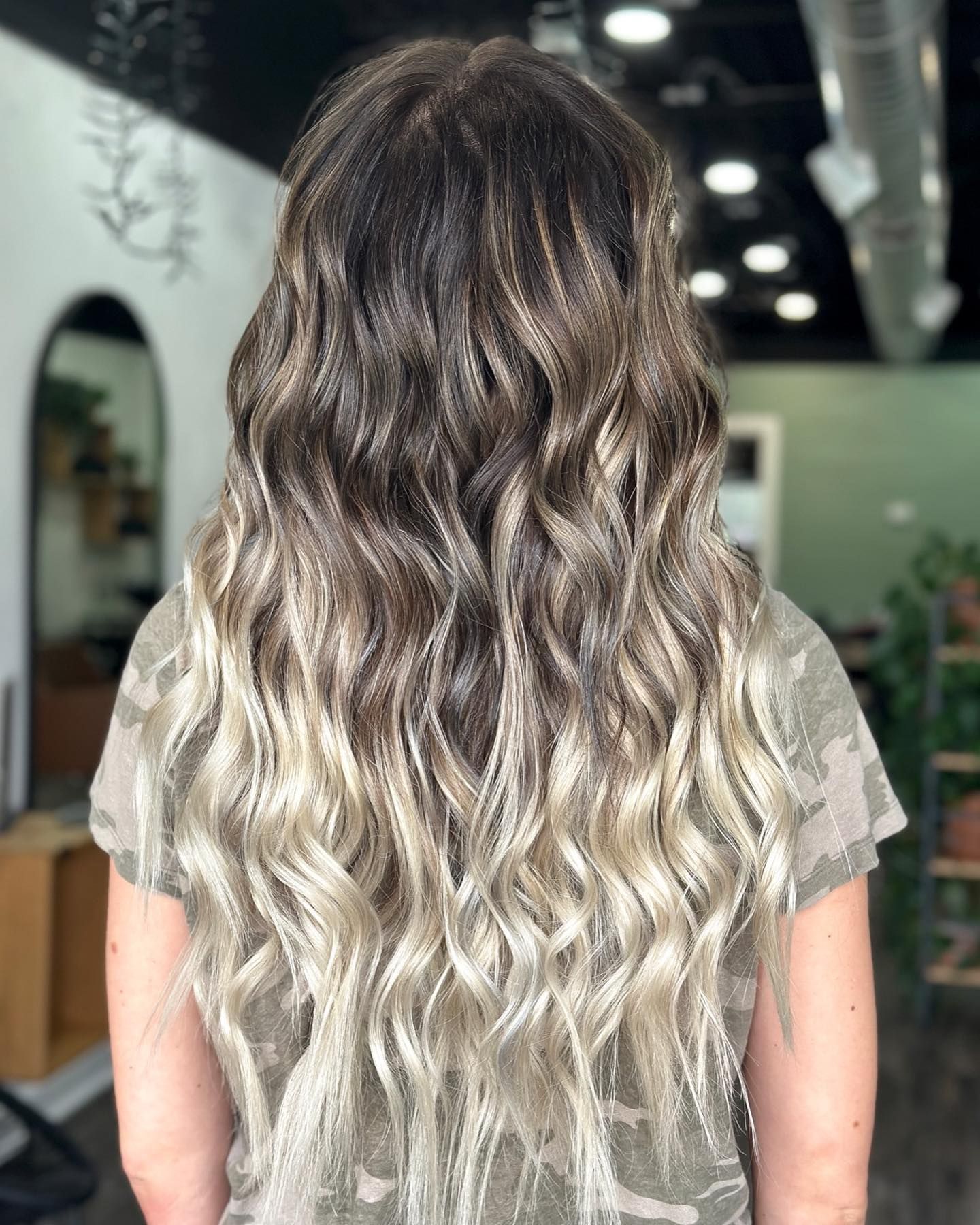 Woman with long wavy hair in a dark to light ombre style, standing inside a salon.