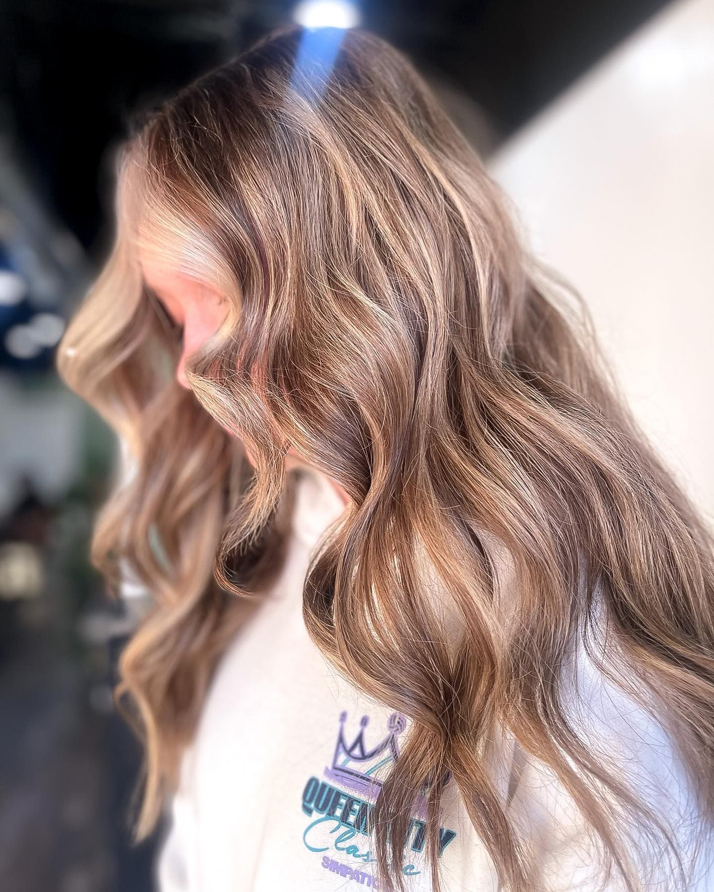 Woman with wavy, highlighted hair wearing a white sweatshirt with a purple logo.