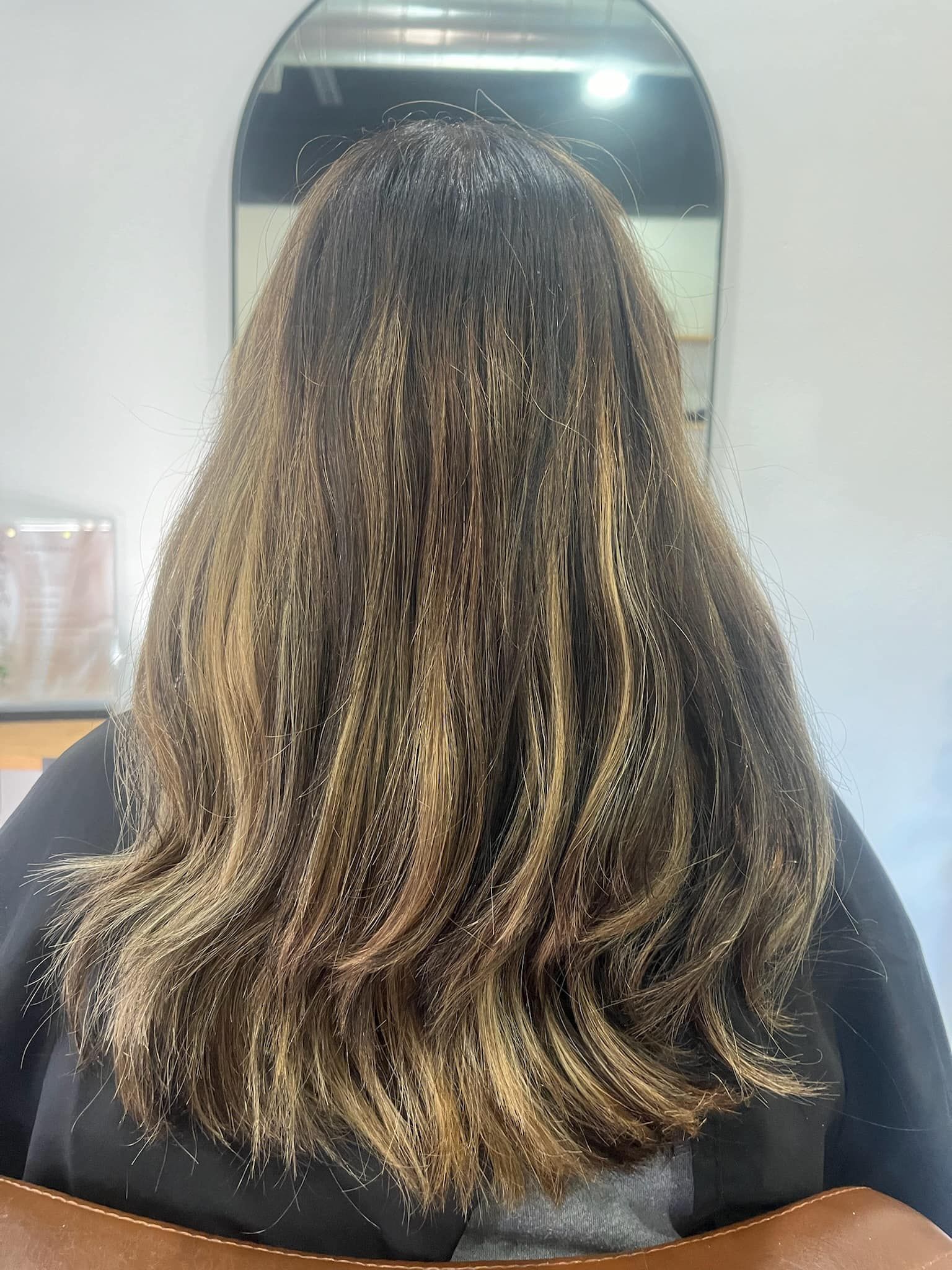 Woman with long, wavy hair; dark roots with light brown highlights. In a salon chair in front of a mirror.