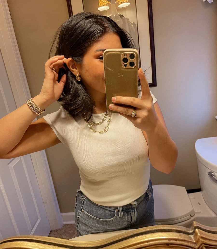 Woman taking a mirror selfie in a bathroom, wearing a white top and holding a gold phone.