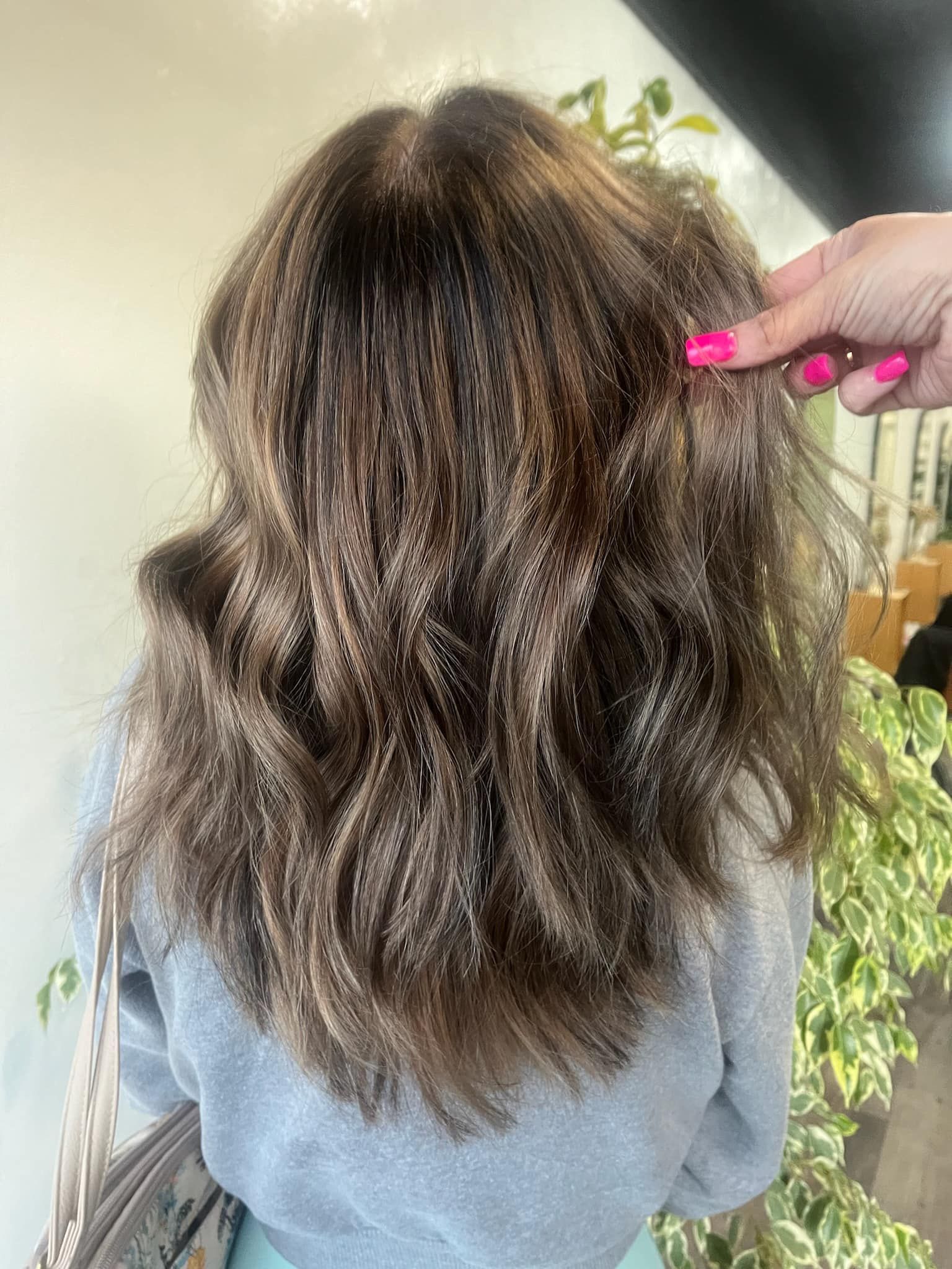 Back view of hair with crimped texture, highlighted brown and blonde. A hand with pink nails touches the hair.