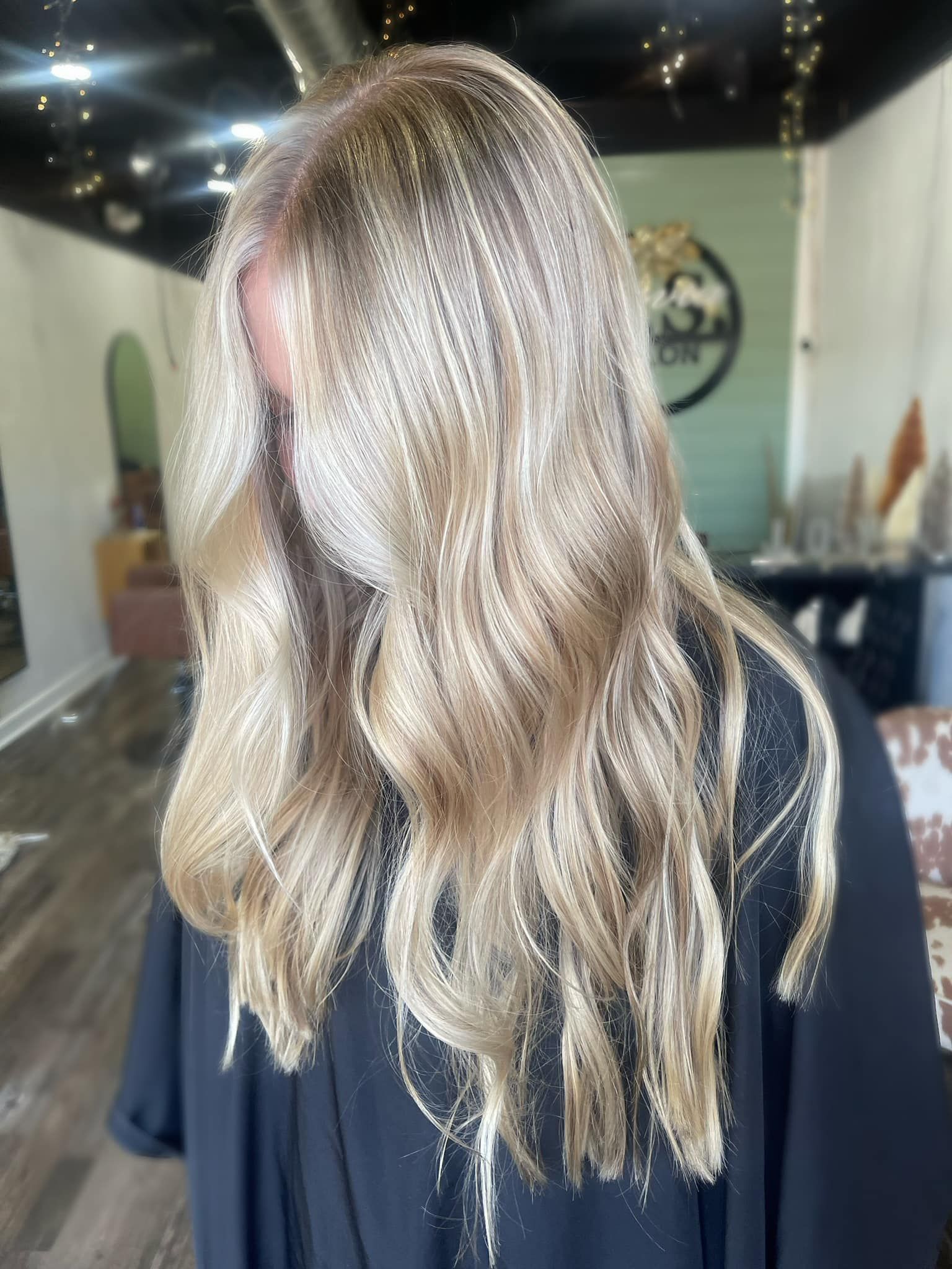 Woman with long, wavy blonde hair wearing a black top and face mask in a salon.