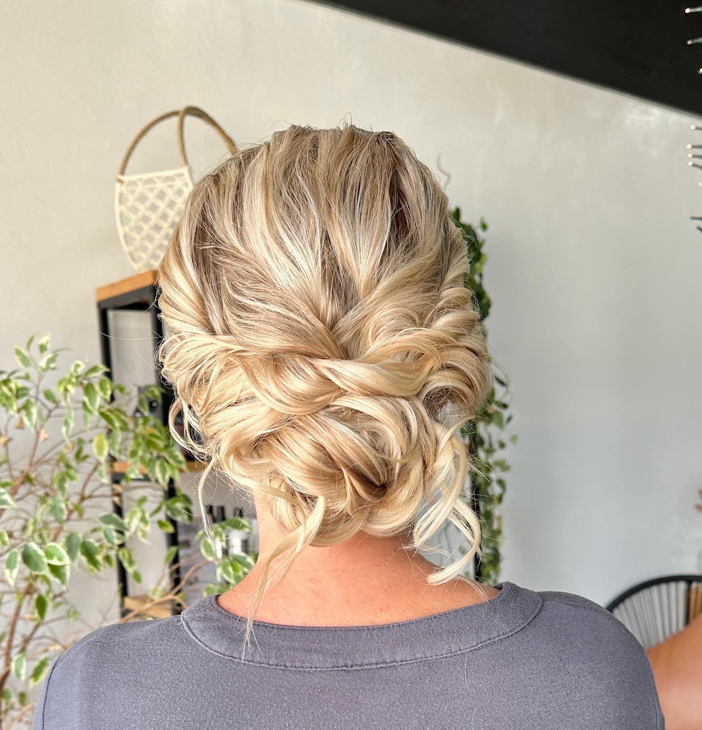 Woman with blonde updo hairstyle, against a light wall, plants visible in background.