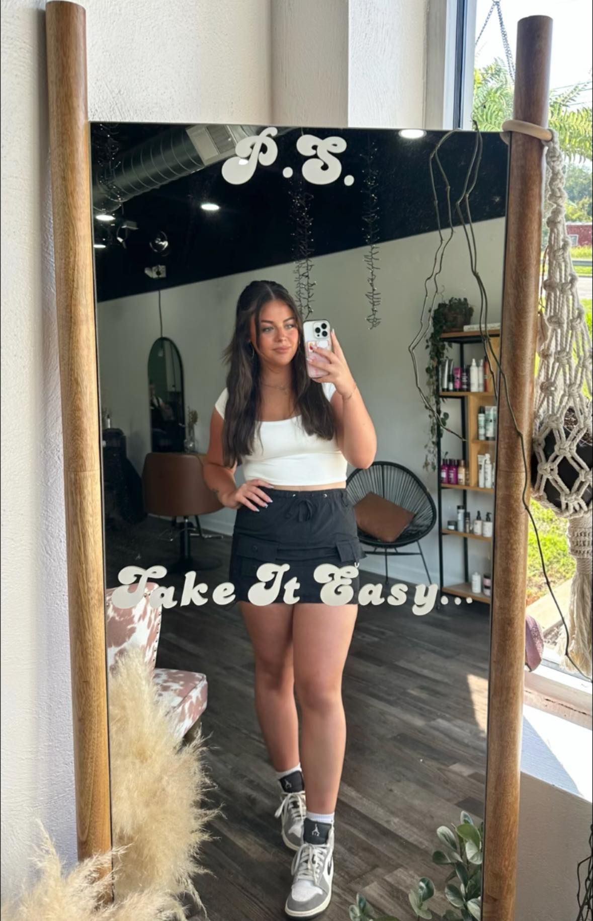 Woman in a white top and black skirt taking a mirror selfie in a salon, holding a phone.