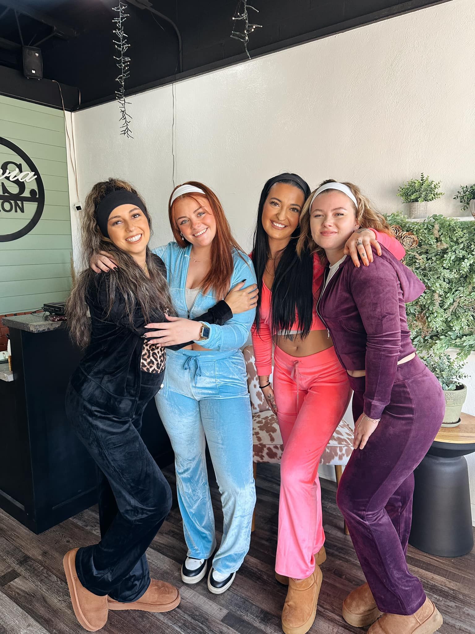 Four women in matching velour tracksuits, posing indoors, smiling.