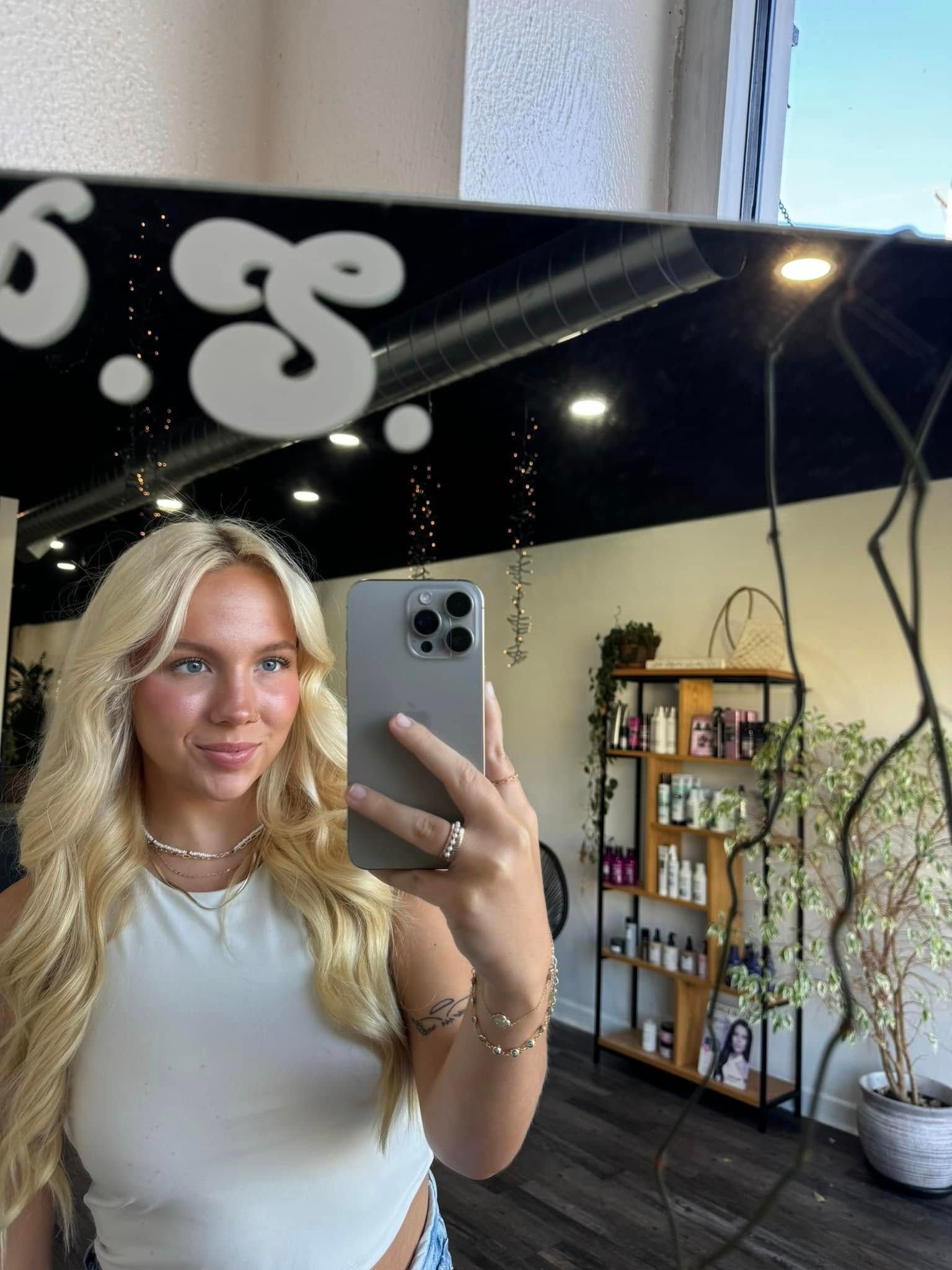 Blonde person taking a mirror selfie in a salon, wearing a white top and jewelry.