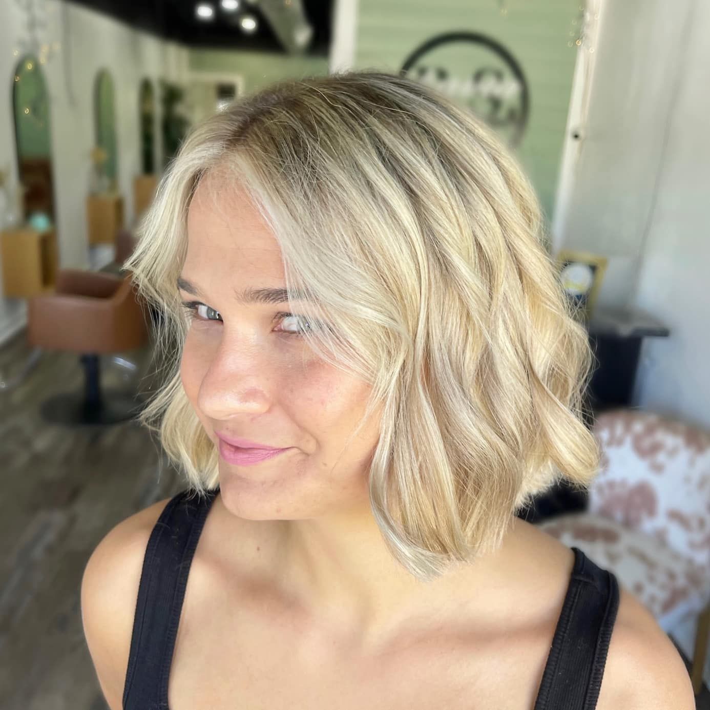 Blonde-haired woman with wavy bob, wearing a black tank top, smiling slightly in a salon.