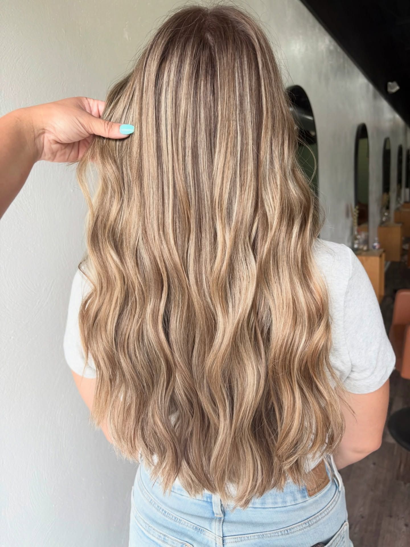 Woman with long, wavy blonde hair with highlights, standing in a salon.