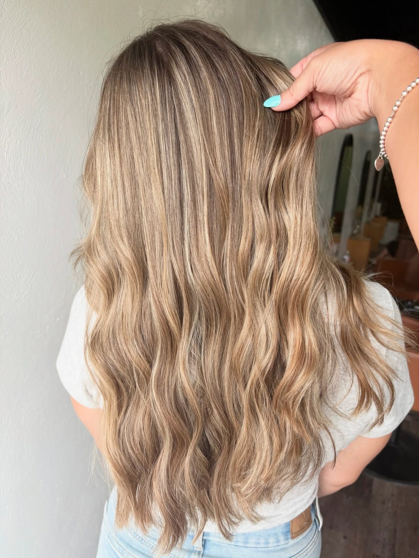 Woman with long, wavy, highlighted brown hair. Hand holding a strand. Light-colored shirt and jeans.