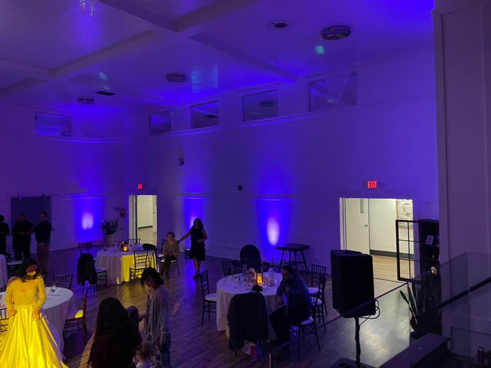 Formal event: Tables set with white linens, black chairs, decorations, and a stage with lights.