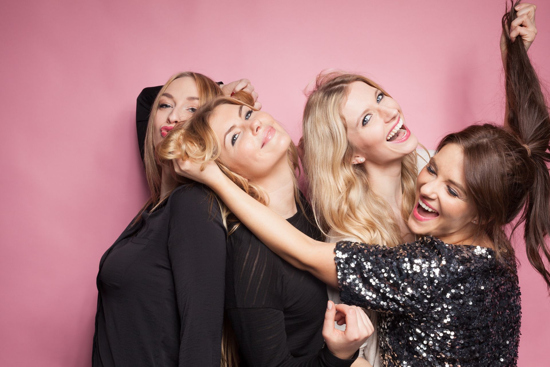 Four smiling women playfully pose; one pulls another's hair, set against a pink backdrop.