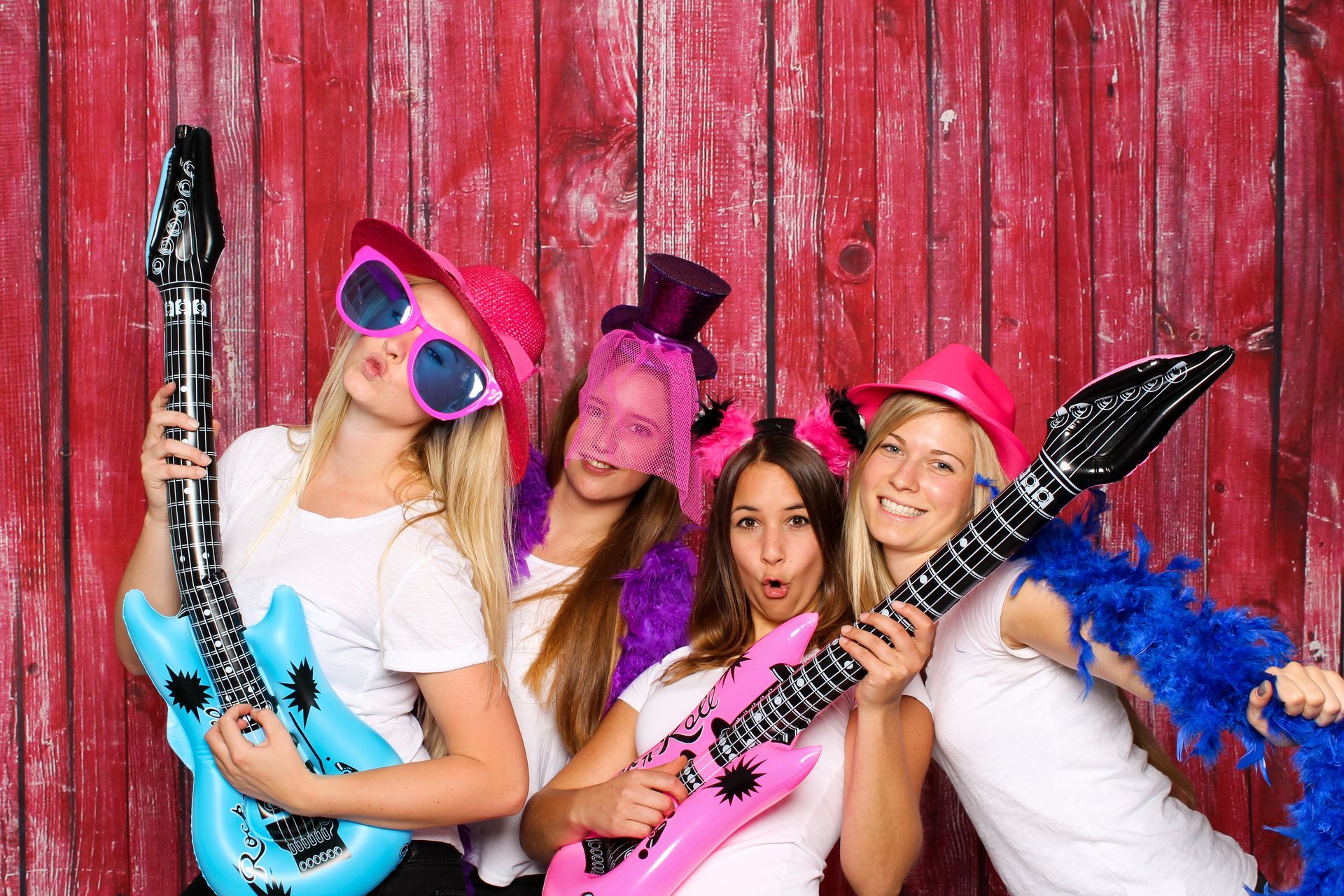 Four people with inflatable guitars and party hats pose in front of a red wooden wall, making silly faces.