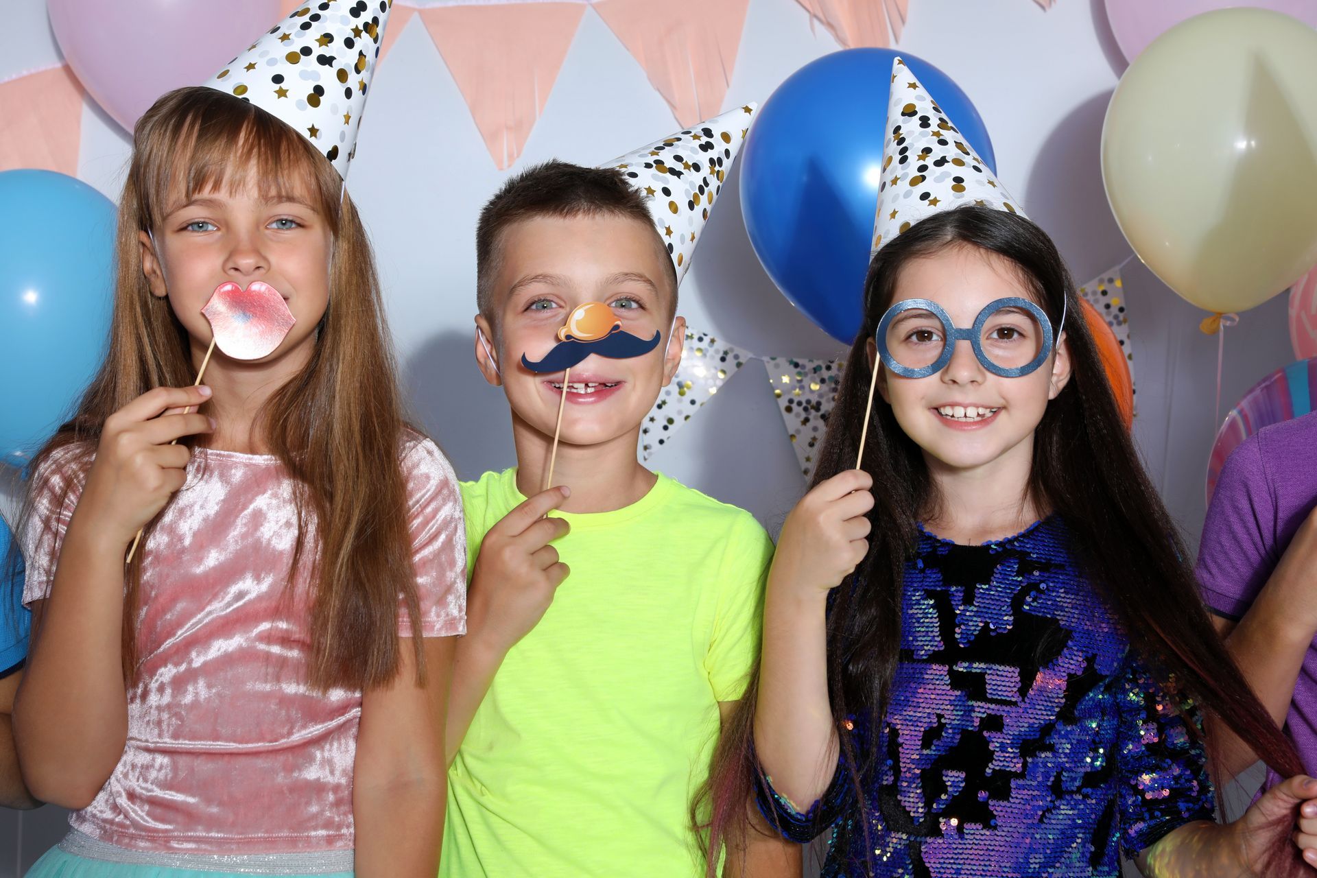 Three children at a party wearing party hats and holding props, smiling at the camera, with balloons and decorations in the background.