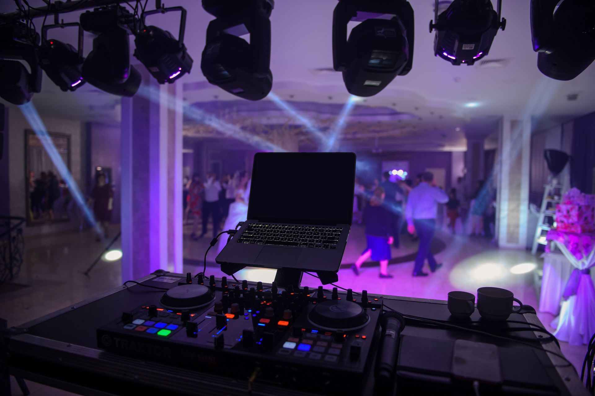 DJ setup with laptop in front of a party; lights and people dancing in the background.