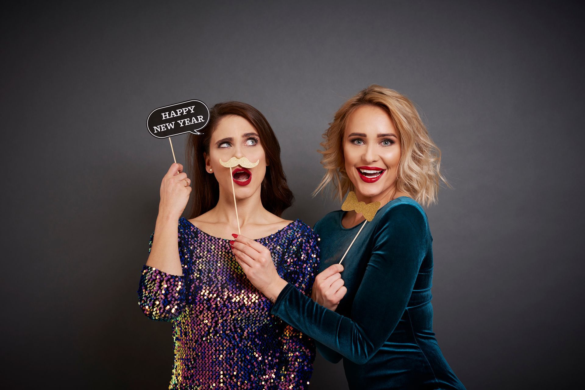 Two women with photo booth props. One makes a surprised face, the other smiles. Against a gray backdrop.