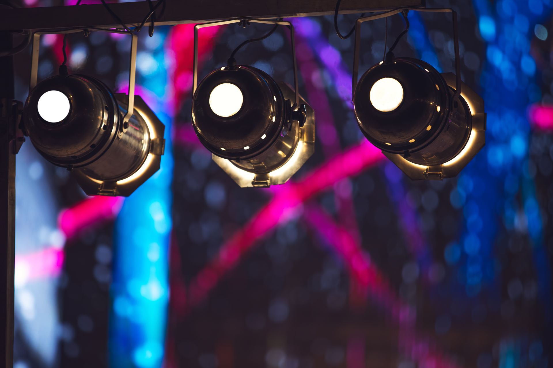 Three spotlights illuminate, against a background of blurred blues, pinks, and purples.