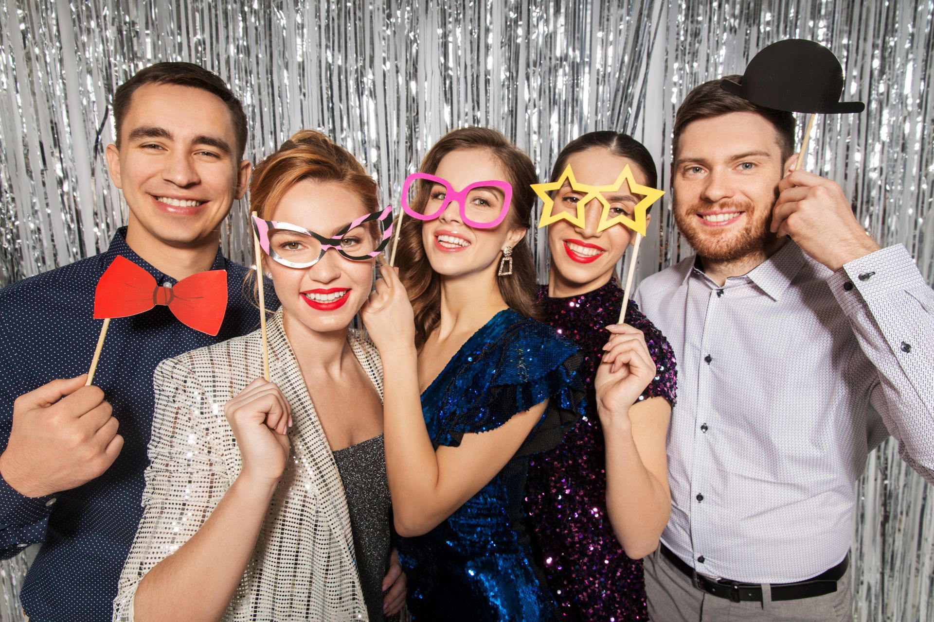 Five people posing with props in front of a silver streamer backdrop. Smiling faces, holding glasses, bow tie, and hat props.