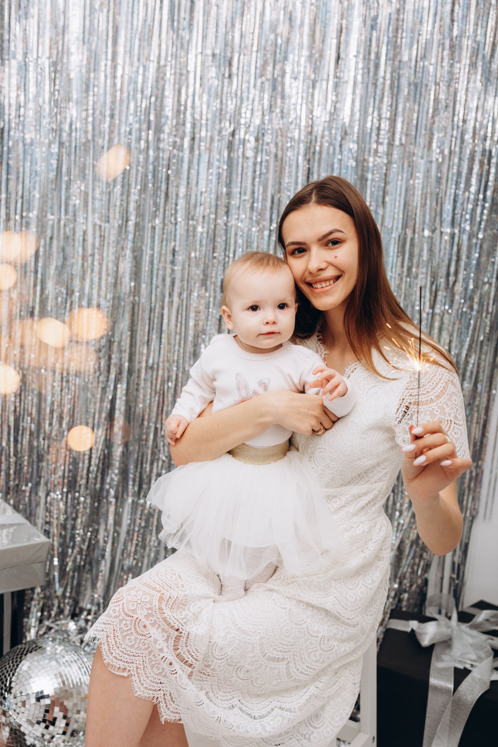 Woman holding baby with sparkler, both in white outfits, in front of silver tinsel backdrop.