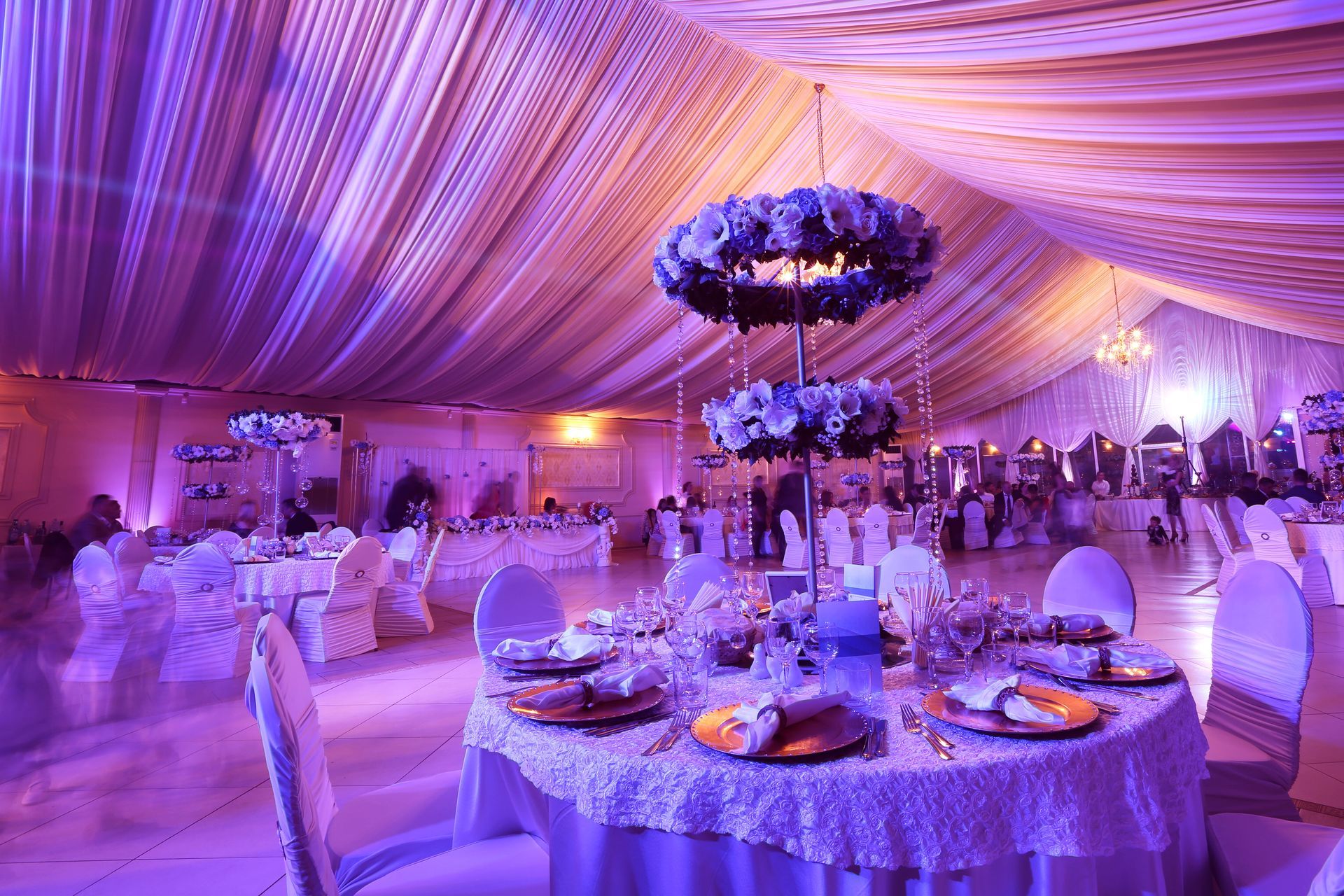 Elegant event hall with draped ceiling, tables set with place settings, floral centerpiece, and purple lighting.