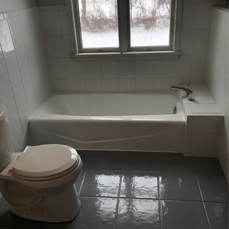 A bathroom with a white tub, toilet, and gray tiled floor. A window is above the tub.