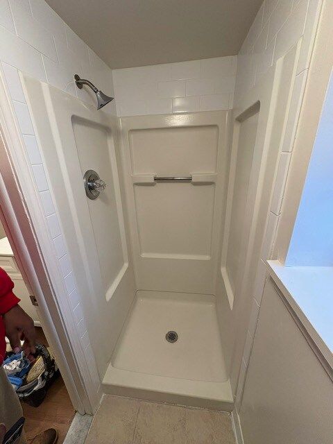 A small, white shower stall with a built-in seat and showerhead. A person holds a tool bag in the bottom left corner.