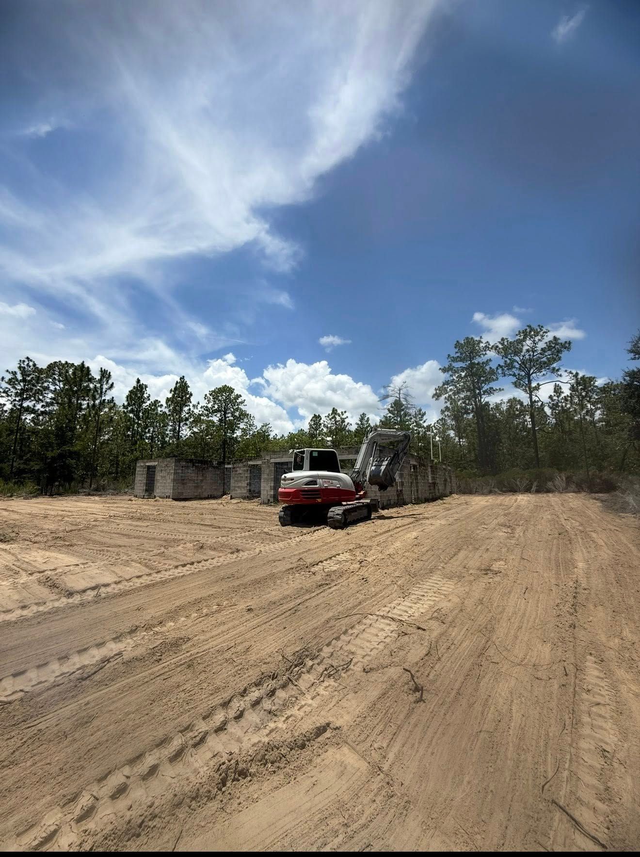 Excavator on a sandy construction site under a blue sky, with trees in the background.