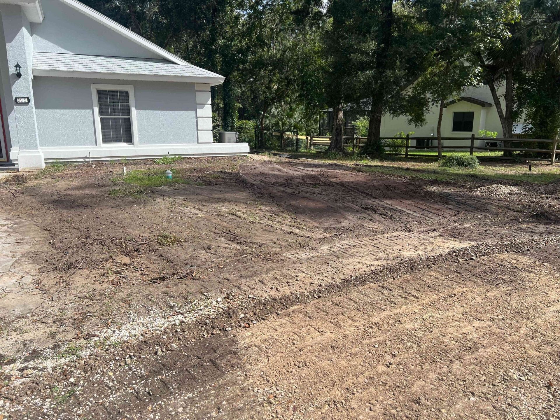 Bare dirt yard in front of a house with a white exterior; a second house and trees are in the background.