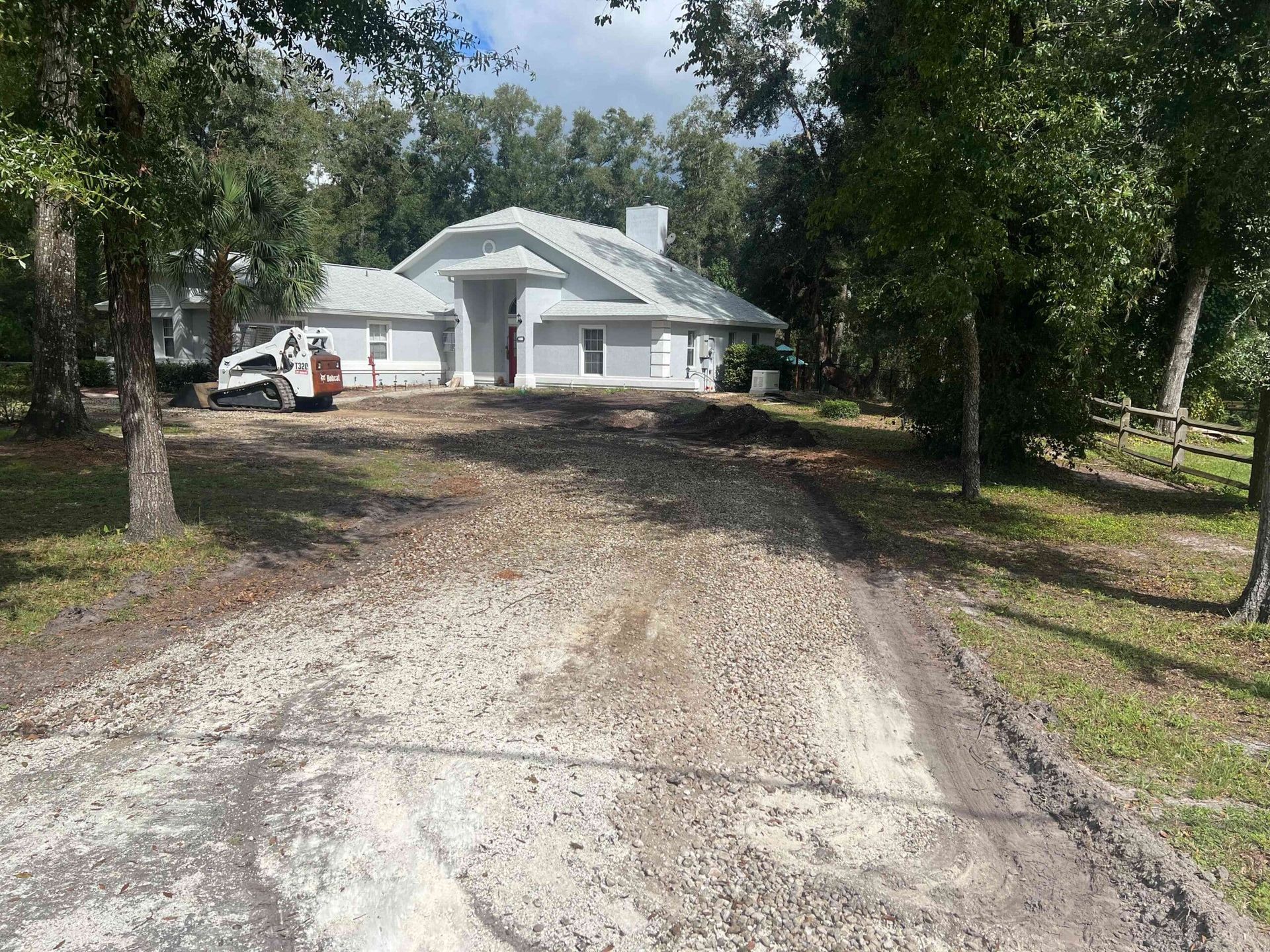 A house with a light gray roof and walls is under construction. A gravel driveway leads to the house.