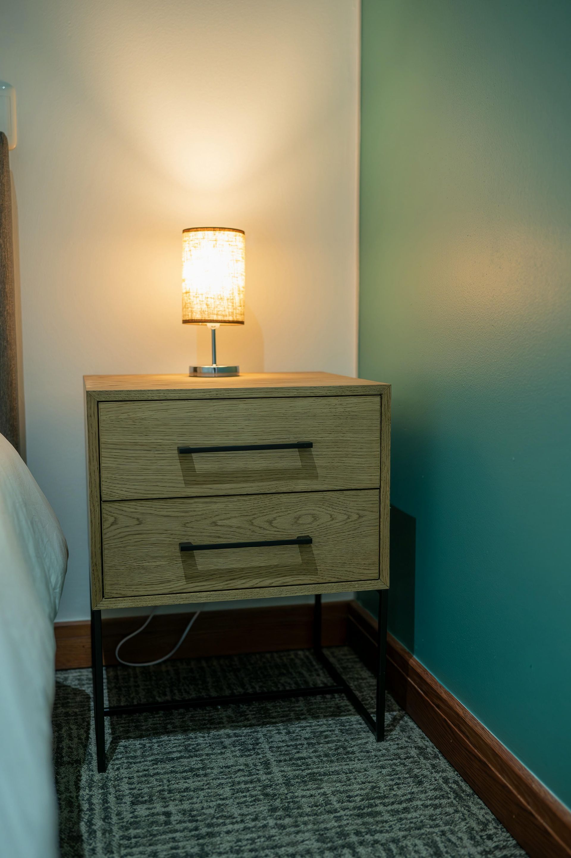 A nightstand with a lamp on it in a bedroom next to a bed.