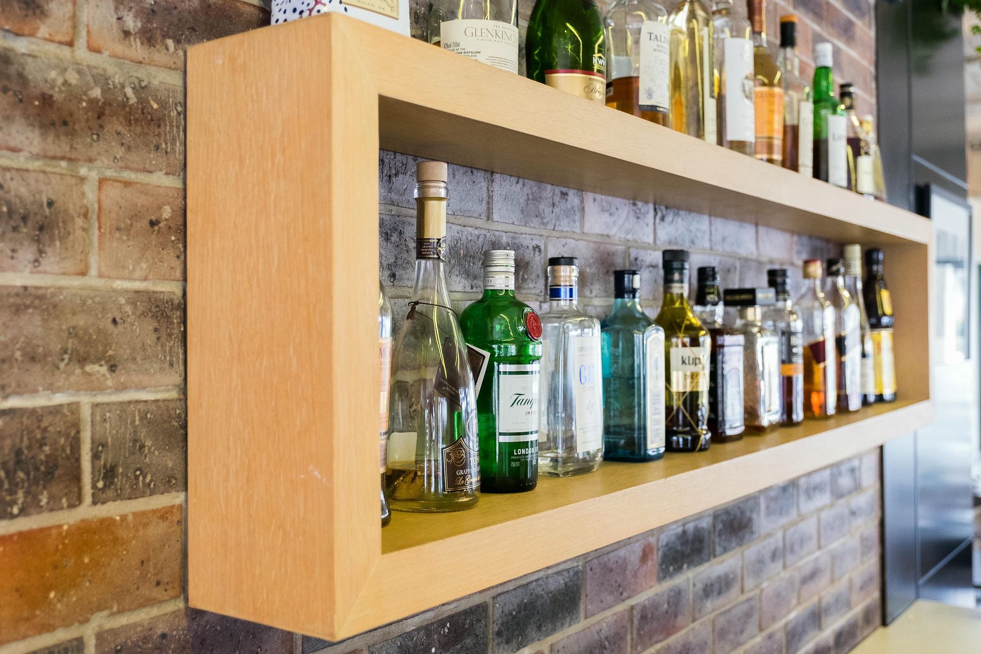 A wooden shelf filled with bottles of alcohol on a brick wall.