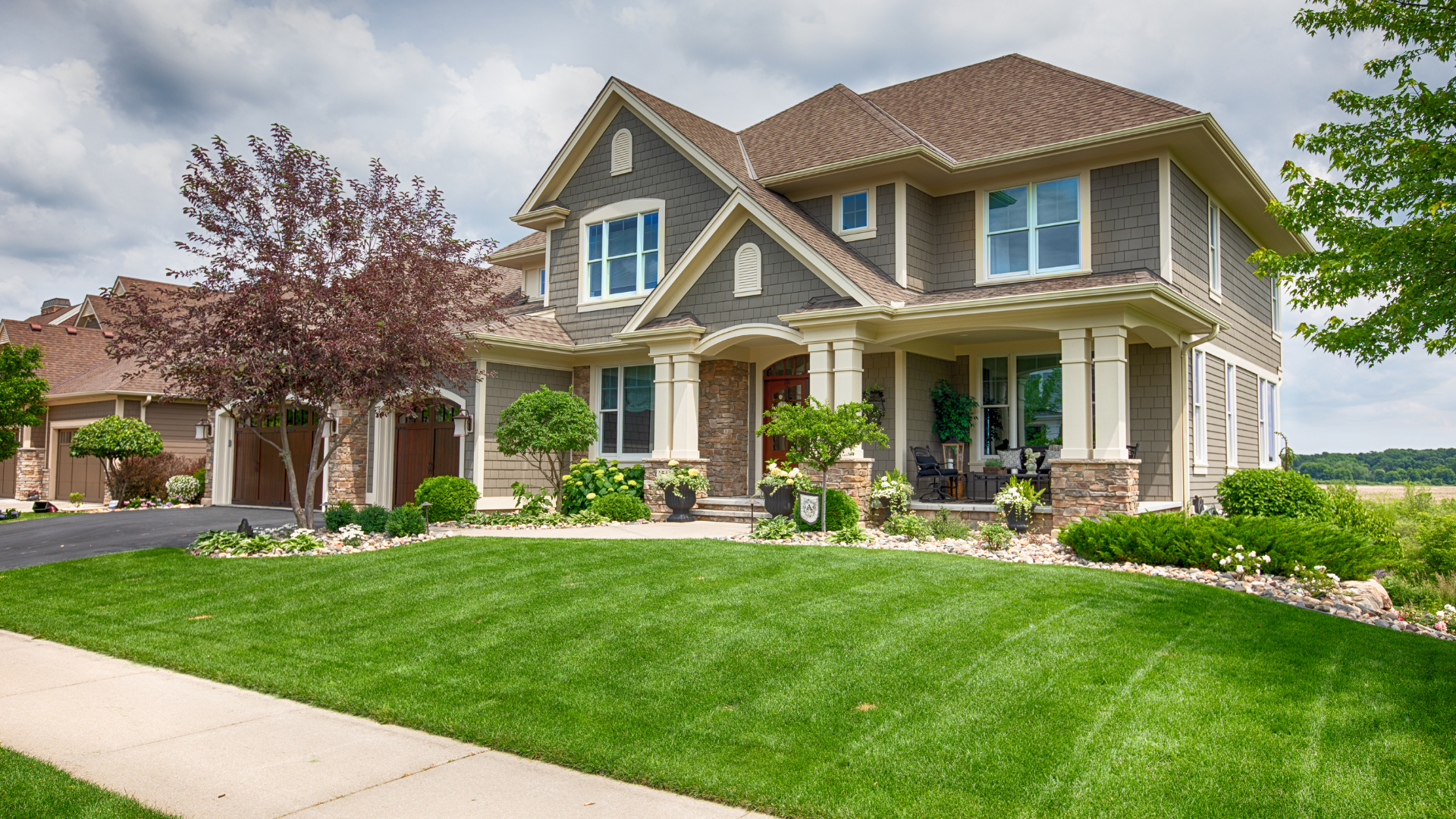 Large suburban house with a manicured green lawn, front porch, and landscaped garden under a cloudy sky