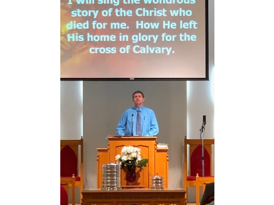 A man stands at a podium in front of a screen that says i will sing the world 's story of the christ