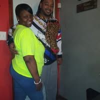 A smiling woman and man pose together. The woman wears a yellow shirt and blue pants, while the man wears a patterned jacket and gray pants. They stand near a door.