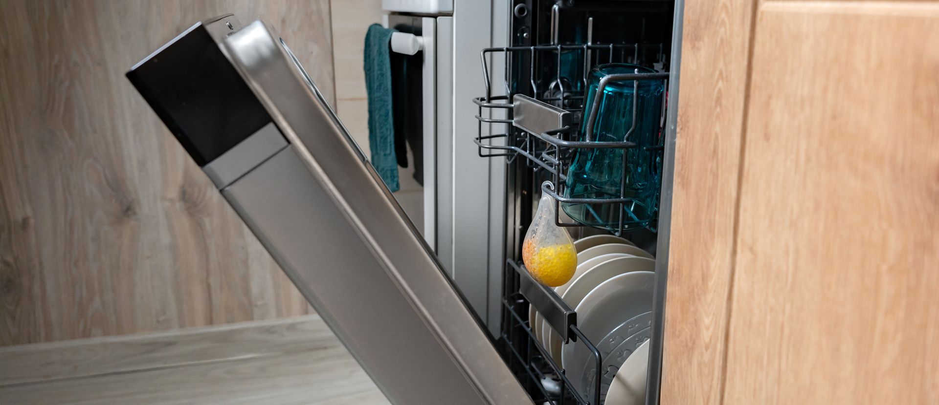 Open dishwasher with light dishes, blue glasses and air freshener on wooden kitchen background. Water-saving and energy-efficient household appliance Open dishwasher with light dishes, blue glasses and air freshener on wooden kitchen background. Water-saving and energy-efficient household appliance