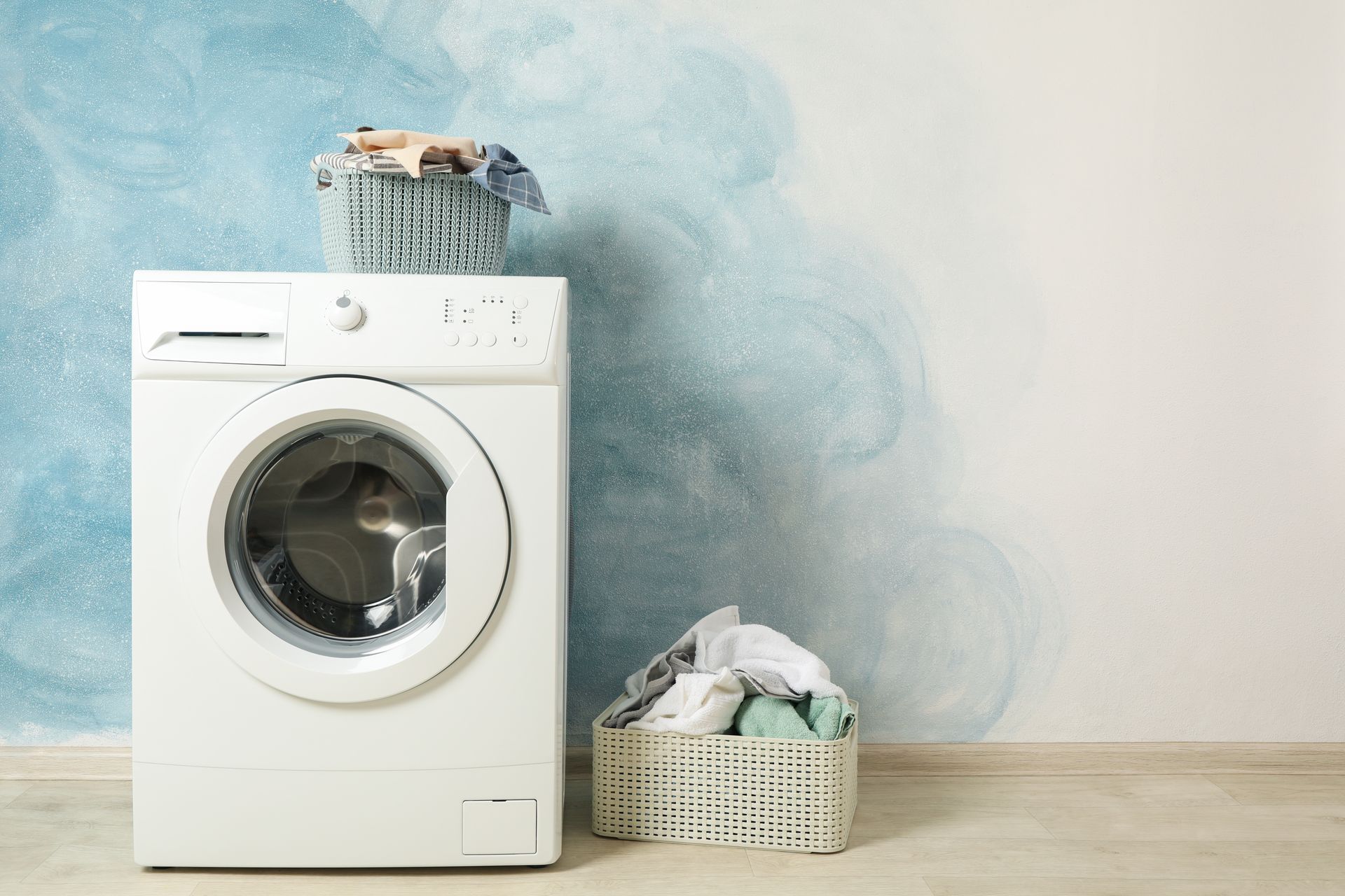 Washing machine with laundry basket on top and a basket of clothes beside it against a blue wall.