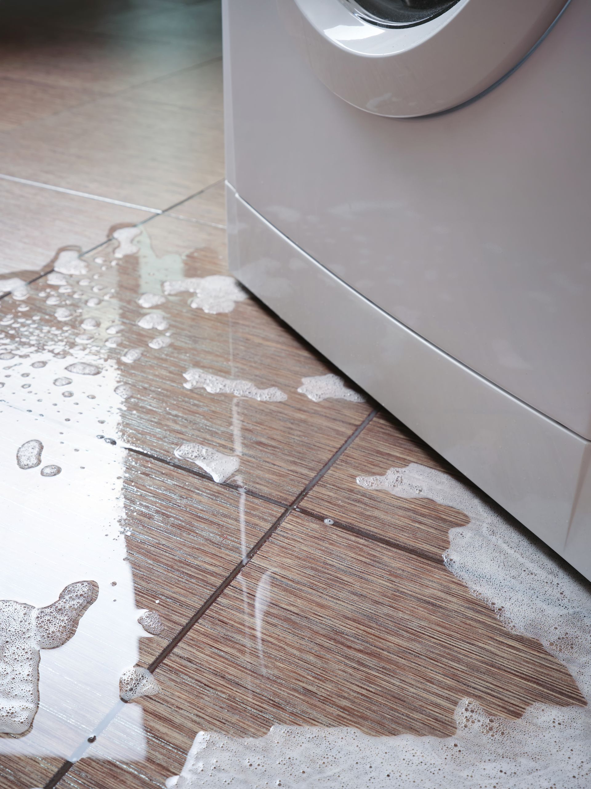 Washing machine leaking water onto a tile floor. White machine, brown and beige tiles.