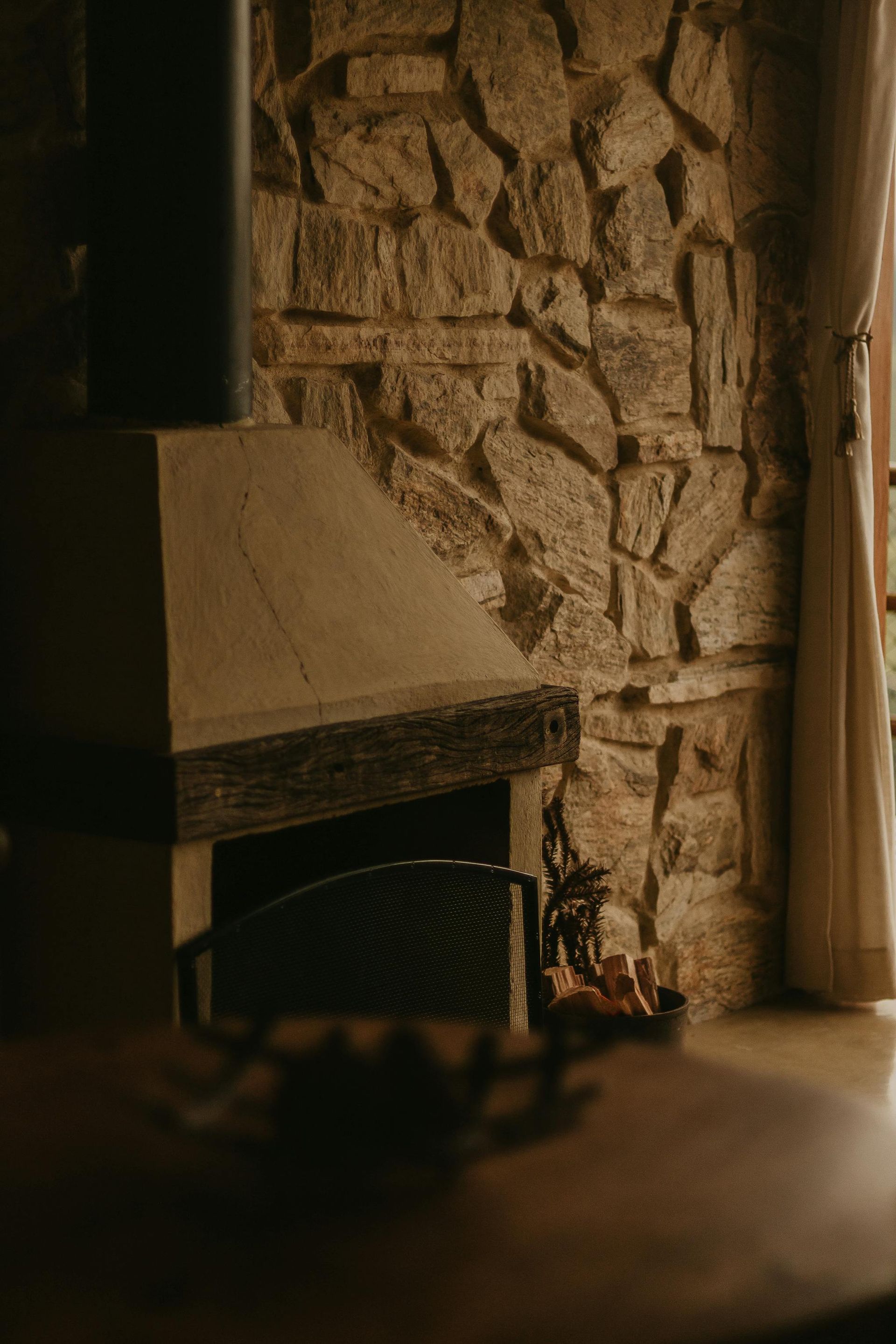 Stone fireplace with black metal screen, wood, and a window with a sheer curtain.