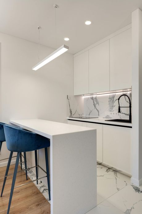 Modern white kitchen with marble backsplash, island with blue bar stools, and linear light fixture.