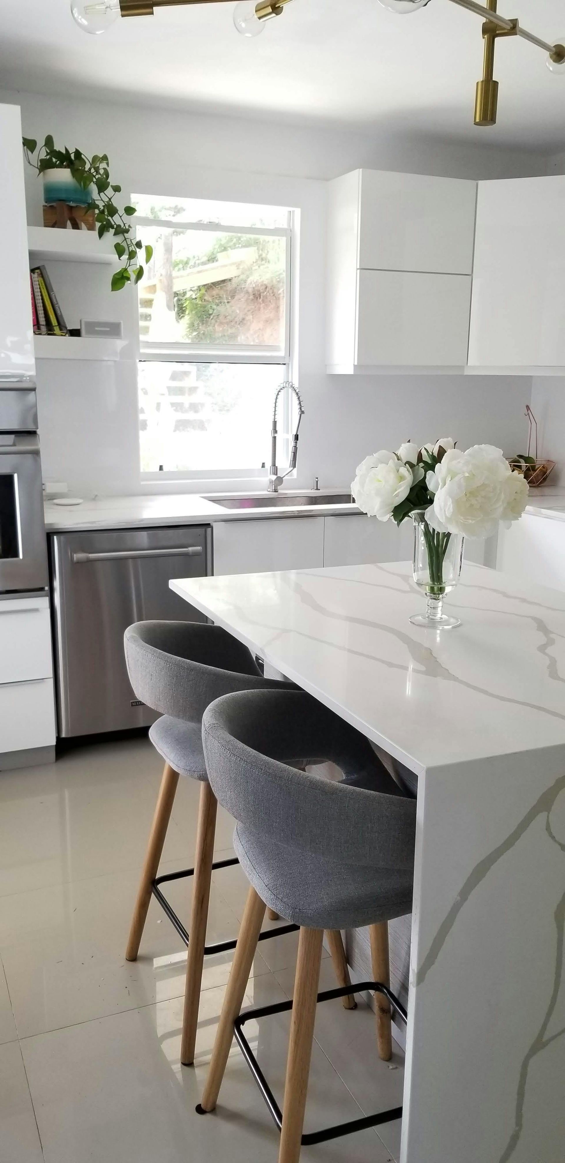 Modern white kitchen with island and bar stools, a window, and a vase of white flowers.