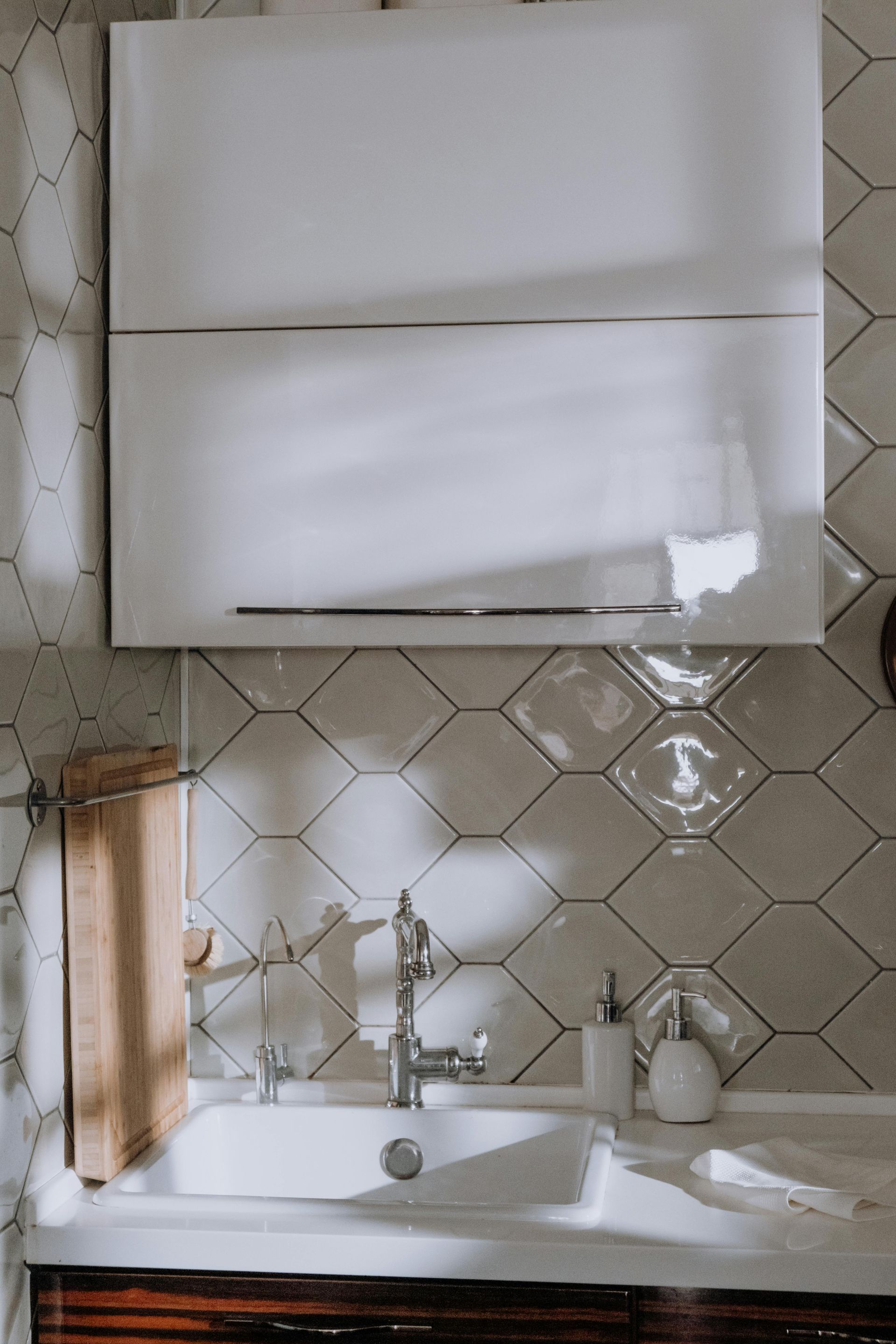 White kitchen sink with faucet, soap dispenser, and cabinetry, set against diamond-patterned tile.