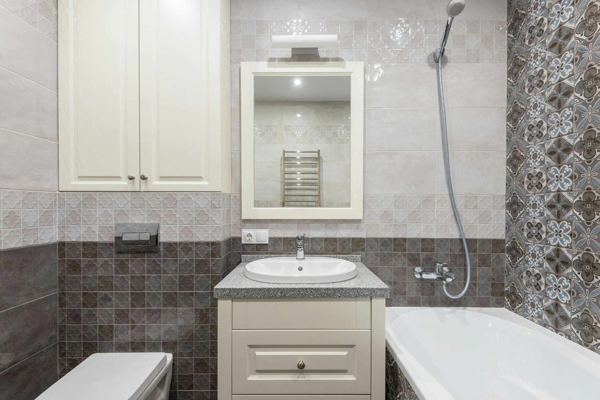 Bathroom with light-colored vanity, mirror, and cabinets, a bathtub, and tiled walls in shades of gray and white.