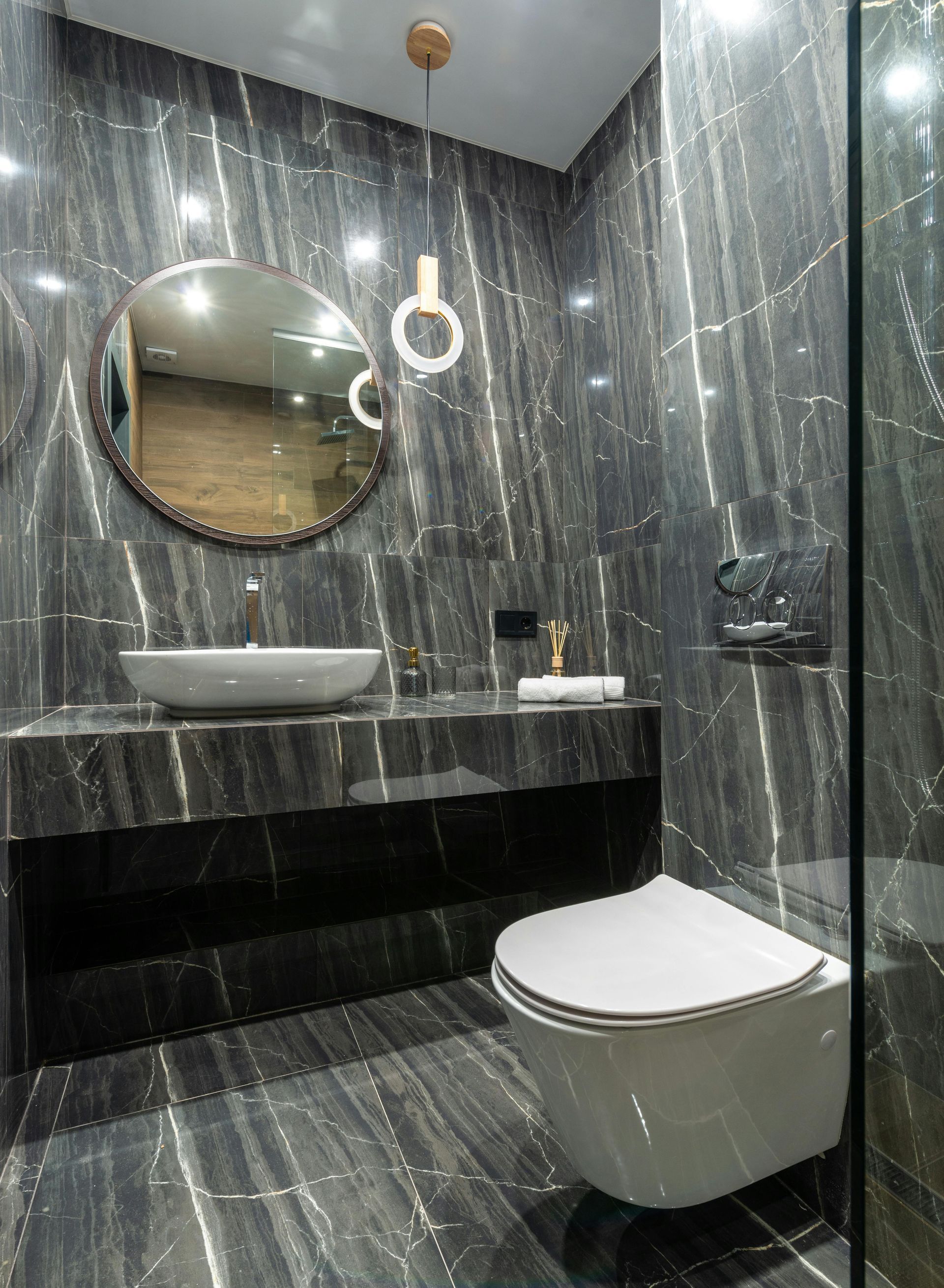 Modern bathroom with black marble walls and floating vanity, round mirror, and a wall-mounted toilet.
