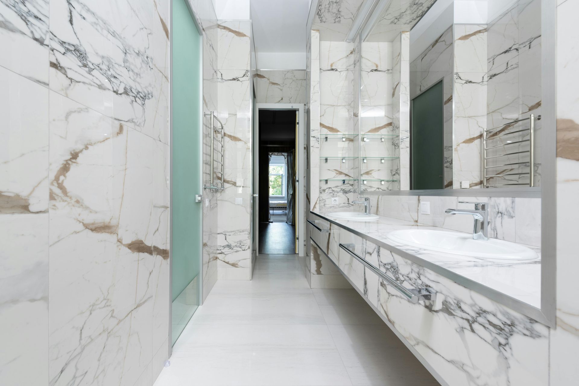 Marble-tiled bathroom with a long counter and two sinks, a glass shower, and a doorway. White and gold veining.