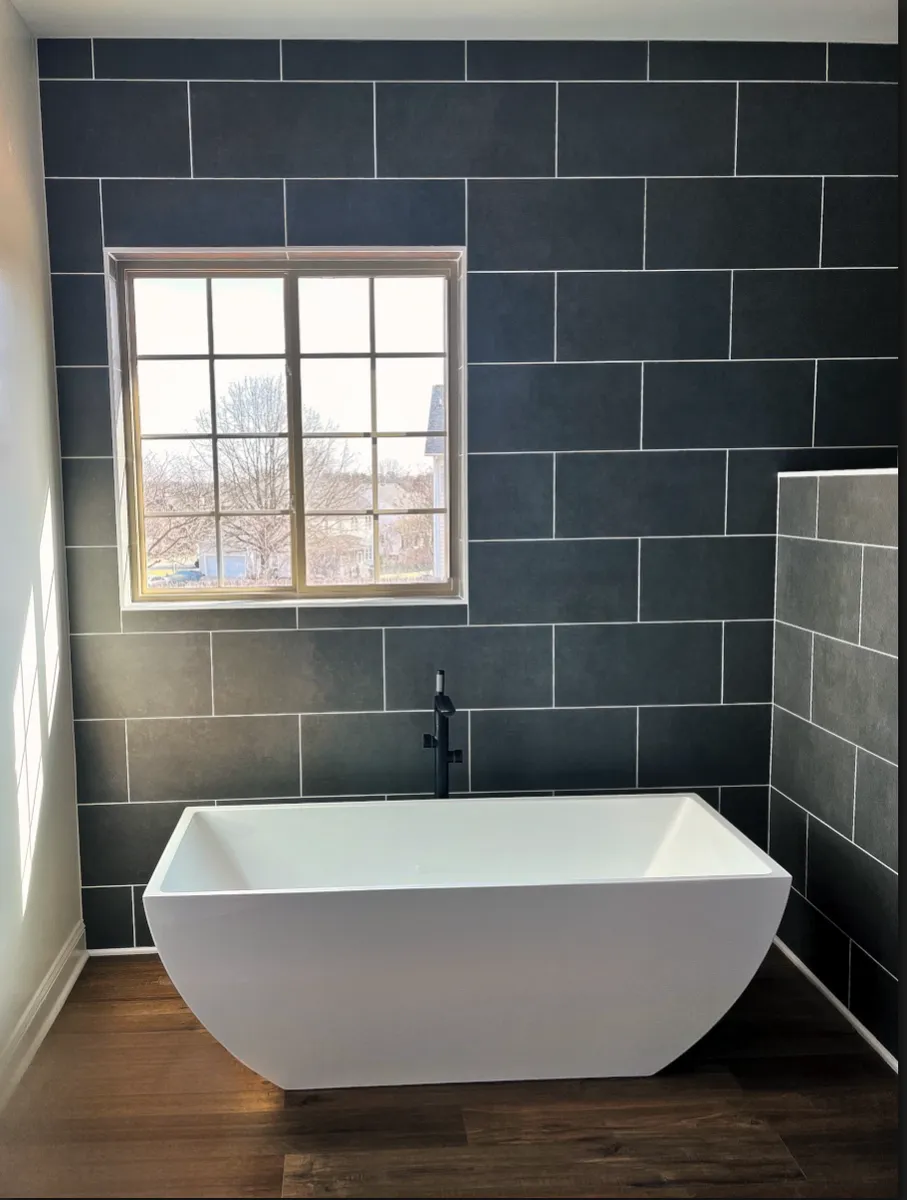 Freestanding white bathtub under a window in a bathroom with dark gray tile walls and wooden floors.