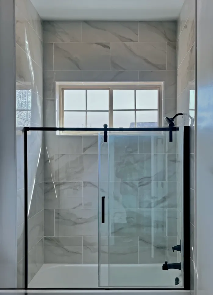 Bathroom with a sliding glass shower door and window. Gray marble-look tiles. Black hardware.