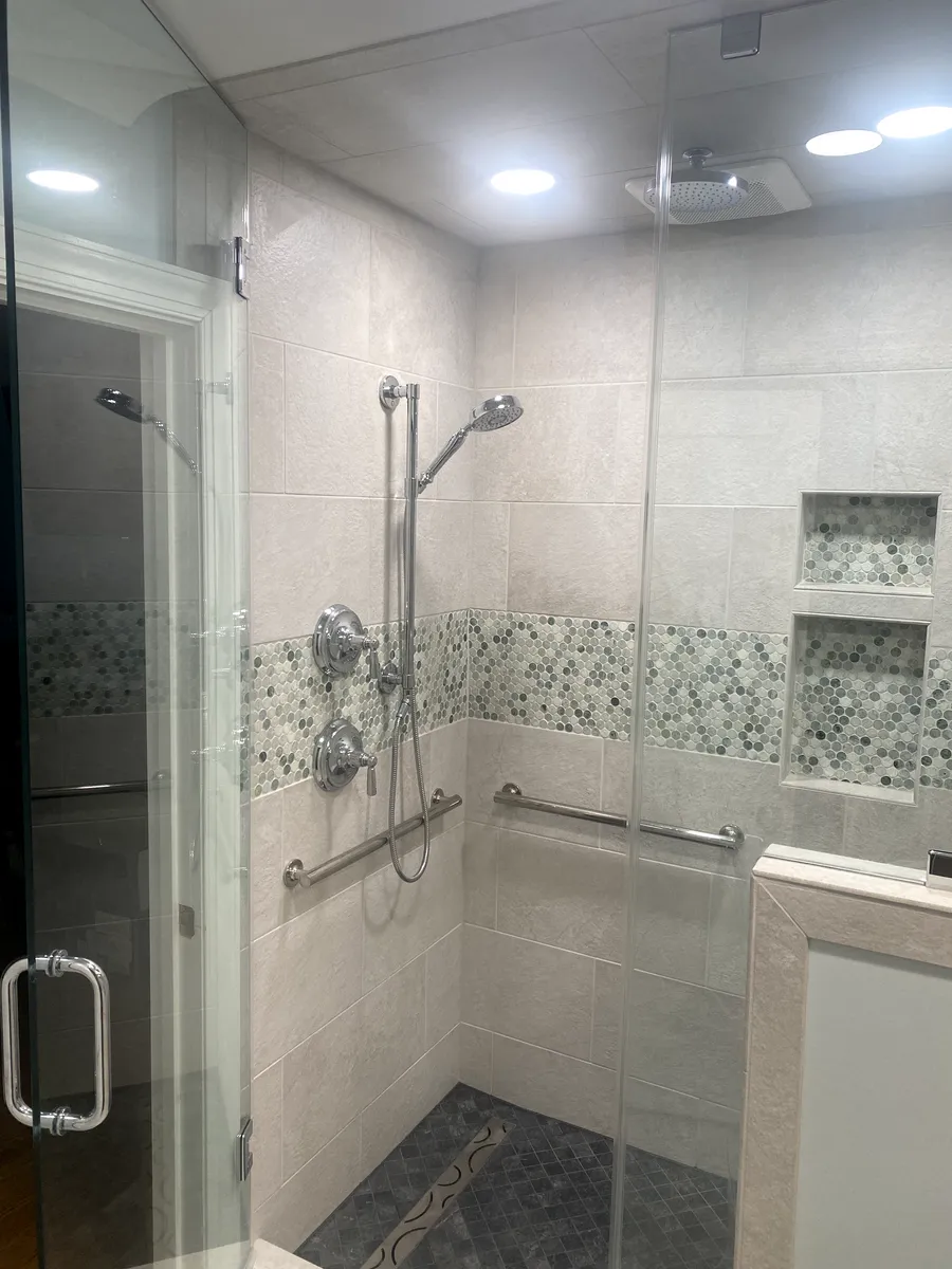 Shower with glass door, grab bars, and neutral-colored tile accented by a decorative band of green and blue stones.