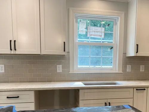 White kitchen cabinets with black handles, light gray tile backsplash, and window.